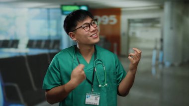 Yeşil önlüklü genç Asyalı bir adam havaalanı terminalinde gitar çalarken şakacı bir şekilde taklit ediyor. Kapalı mekan ortamında samimi ve komik bir ifade sergiliyor..