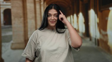 Genç ve cüsseli bir kadın, eski üniversite koridorunda kemerli, kendine güvenen ve günlük tarzını sergileyen jest yapıyor..