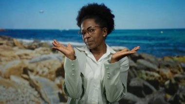 Deniz kenarında güvenle duran, arkasında kayalar olan bir kadın, açık mavi gökyüzünün altında zarif bir varlığı tasvir ediyor, açık havada gözlük takıyor..