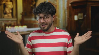 Sakallı ve gözlüklü genç adam çizgili gömlek giyiyor. Arka planda mumlar ve heykellerle kilisenin içinde sürpriz hareketler yapıyor..