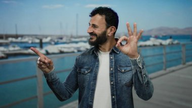 Sakallı İspanyol bir adam tekneli bir limanda ve arka planda açık mavi bir denizde rahat rahat giyinip neşeli bir hava yayıyor..