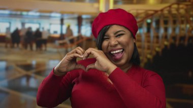 Latin bir kadın otel lobisinde kırmızı giyiniyor, gülümsüyor ve kalp hareketi yapıyor, sıcak, davetkar bir ortam yaratıyor..