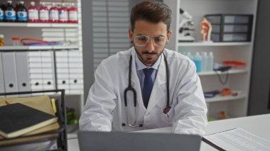Yakışıklı İspanyol erkek doktor hastanede dizüstü bilgisayarla çalışıyor. Steteskopla klinik ortamda beyaz önlük giyiyor. Profesyonel odaklanma ve kendini adamışlık gösteriyor.
