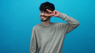 Genç ve yakışıklı bir adamın portresi gülümseyen ve izole edilmiş mavi bir arka plan üzerinde barış işareti yapan, kendine güveni ve arkadaşlığı şık bir kıyafetle yayan.