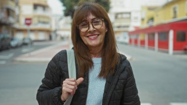 Şehrin sokak ortamında güvenle gülümseyen İspanyol asıllı son sınıf öğrencisi kadın, şehir arka planında, açık havada yakalanmış, açık renk süveter, gözlük ve gündelik ceket, somut modern yaşam tarzı..