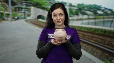 Tren raylarının yanında domuz kumbarası tutan bir kadın gönüllü tişörtü giyiyor. Açık havada huzuru ve amacı vurguluyor.