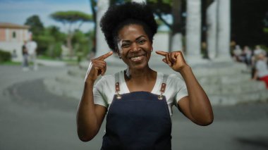 Önlüklü bir kadın açık havada gülümsüyor, bir şehir caddesinde yanaklarına dokunuyor, pozitiflik ve neşe saçıyor, şehir yaşamını ve açık hava ruhunu somutlaştırıyor..