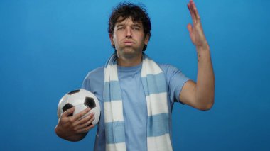 İspanyol adam futbol topunu tutuyor mavi atkı takıyor ve mavi arka planda spor hevesini tasvir eden kararlı bir ifade sergiliyor..