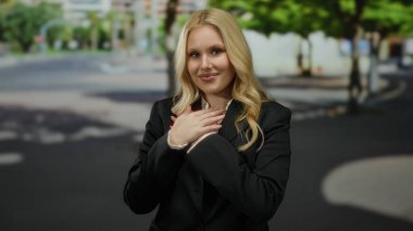 Şehirde minnettarlığını ifade eden bir kadın ağaçlar ve binalar, sarı saçlar, göğsünde eller, siyah bir takım elbise, güneşli bir gün, huzurlu bir atmosfer, gülümseme, dingin bir ruh hali.