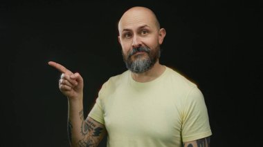 Stüdyoda siyah arkaplanı işaret eden dövmeli ve sakallı bir adam odaklanma ya da karar verme konseptini belirliyor.