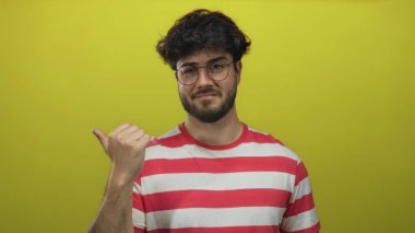 Parmakla işaret eden gözlüklü genç adam, parlak sarı arka planda çizgili gömlek giyiyor, rahat bir ifade ve oyuncu bir jest sergiliyor..