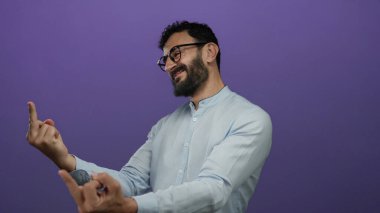 Sakallı ve gözlüklü bir adam mor bir arka planda duruyor. Cesur bir el hareketi yaparken açık mavi gömlekli, etkileyici duruşlu bir şekilde gülümsüyor..