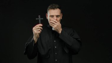 Rahip gibi giyinmiş bir adam karanlık bir arka plana karşı elinde haç, ağzının üzerinde bir el, sürpriz ya da şok, izole edilmiş ve gizemli dini imgeler iletiyor..