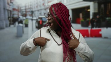 Afro-Amerikalı bir kadın, hareketli bir şehir caddesinde hareketli bir şehir manzarası sergileyerek kendine sürpriz yüz ifadeleri sergiliyor..