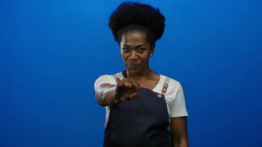 Önlük takan genç bir kadın, izole edilmiş mavi arkaplan karşısında şakacı hareketler sergiliyor.