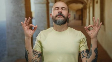Eski bir üniversitede meditasyon yapan bir adam gözleri kapalı ve dövmeleri görünür, sakin ve huzurlu bir atmosfer yaratıyor..