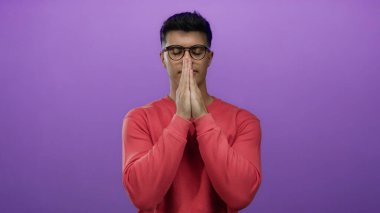Kırmızı kazaklı genç adam mor bir arka plana karşı dua ediyor, elleri bir arada, gözleri kapalı, izole edilmiş, sessiz iç gözlem ifade ediyor..
