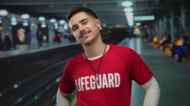 Bıyıklı ve küpeli genç İspanyol adam can kurtaran tişörtü giyiyor. Güvenli bir şekilde tren istasyonunda duruyor. Arka planda parlak ışıklar ve platform görünüyor..