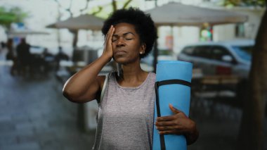 Şehir caddesindeki terasta dinlenen bir kadın yoga minderini kapalı gözlerle tutuyor, şehir hayatının ortasında sükuneti somutlaştırıyor, bulanık bir restoran geçmişine karşı günlük giysiler giyiyor..