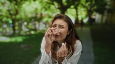 Neşeli yüz ifadeleri olan genç bir kadın, yeşil ve ağaçlarla çevrili güneşli bir parkta parmaklarını çimdikleyerek komik bir an yaşatıyor..