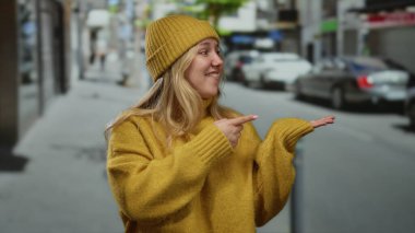 Sarı kazak ve şapkalı bir kadın, bir şehir caddesini işaret ediyor, canlı bir şehir manzarasında şehir yaşamını gözler önüne seriyor..