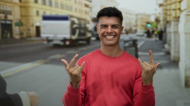 Kırmızı süveter giyen genç adam güler yüzlü ve hareketli bir şehir caddesinde gün ışığında coşkuyla el kol geziyor..