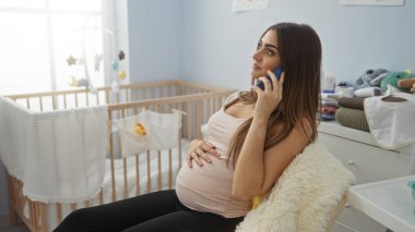 Hamile kadın telefonda konuşuyor, rahat bir bebek odasında, sandalyesinde bir beşiğin yanında, yumuşak ışıklandırma ve bebek dekoruyla, sakin bir beklenti ve hazırlık iletiyor..