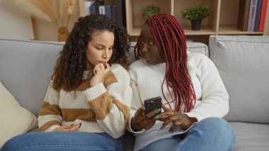 Two women friends sitting indoors on a cozy sofa in a modern apartment discussing something intriguing while one holds a smartphone and the other looks curious and engaged