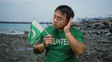 Plajda elinde Pakistan bayrağı olan genç bir adam dikkatle dinliyormuş gibi kulak kesiliyor, yeşil gönüllü gömleği giyiyor, deniz kenarındaki kültürel çeşitliliği temsil ediyor..