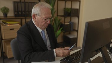 Akıllı telefon kullanan kıdemli bir adam modern ofiste masasında oturuyor ve başarılı haberlerin heyecanını ve neşesini gösteriyor.