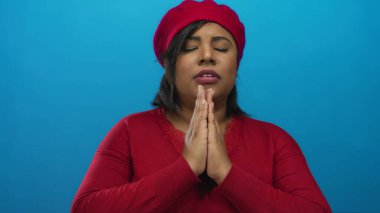 Kırmızı bereli ve kazaklı bir kadın, canlı mavi bir arka planda duruyor, umudunu ve inancını kapalı gözlerle ifade ediyor..