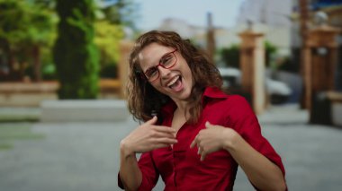 Kırmızı tişörtlü kadın şehir sokaklarında ağaçlar ve binalarla mutlu bir şekilde el kol hareketi yapıyor, gözlük takıyor, neşeli bir ifade sergiliyor..