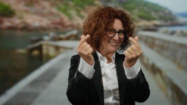 İspanyol kadın deniz kenarında gezinirken parmaklarıyla kalp hareketi yapıyor, siyah bir takım elbise ve gözlük takıyor..
