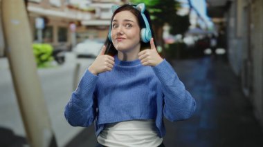 Esmer saçlı, kulaklık takan bir kadın sokak ortasında neşeli bir ifade sergiliyor.