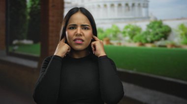 Latin kökenli genç bir kadın dışarıda kulaklarını kapatıyor. Kule 'nin sokak manzarası sıkıntılı görünüyor..