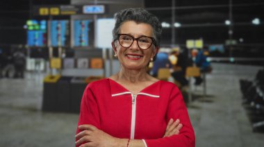 Havaalanı terminalinde kollarını kavuşturmuş kıdemli kadın uçuş bilgileri ve yolcuların bulanık geçmişiyle güvenle gülümsüyor..