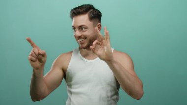 Beyaz askılı sakallı genç adam yeşil bir arka plana gülümseyen, dışa dönük, dışa dönük yüz ifadelerini sergileyen iyi bir işaret veriyor..