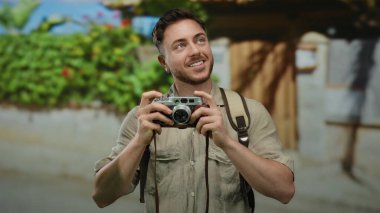 Elinde kamera ve sırt çantası olan beyaz sakallı genç bir adam, canlı bir şehir caddesinde fotoğraf yolculuğunun tadını çıkarıyor..