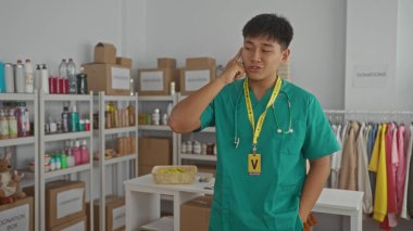 Yeşil önlüklü genç adam, bağış merkezinde telefonla konuşuyor. Etiketli kutular ve giysi rafları var. Gönüllü çalışma ortamını gösteriyor..