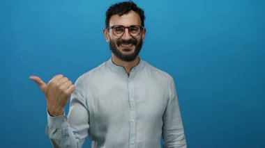 Sakallı, gülümseyen, kendine güveni olan, parlak mavi bir arka planı olan, gözlüklü, açık renk tişörtlü, pozitif ve anlamlı bir duruşu olan İspanyol bir adam.