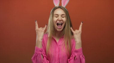 Paskalya tavşanı kulağı takan ve iki eliyle kaya jesti yapan bir kadın, izole edilmiş kırmızı bir arka plana yaslanmış, heyecan ve neşe ifade ediyor..