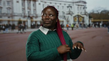 Pembe örgülü bir kadın, tarihi bir bina ve arka planda hareketli bir kalabalıkla güvenli bir şekilde gülümseyerek, canlı bir şehir atmosferini yakalıyor..