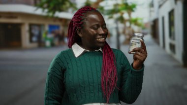 Afrika kökenli Amerikalı bir kadın, elinde bir kavanozla kalabalık bir caddede yeşil bir kazak giyiyor ve düşünceli bir ifade sergiliyor..