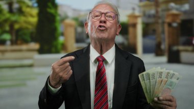 Son sınıf öğrencisi elinde Czech Koruna banknotları tutarak şevkini ifade ediyor, zenginliğini sergiliyor, yeşillik ve bulanık şehir geçmişi olan bir iş ortamında yer alıyor..