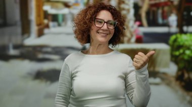Orta yaşlı İspanyol kadın, şehir sokaklarında sırıtan gözlüklü, kıvırcık esmer saçlı, sıradan giyinen ve pozitifliğini baş parmaklarıyla ifade eden bir kadın..