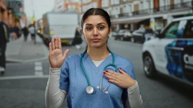 İspanyol kadın doktor yemin jesti yapıyor. Bir eli yüreğinde, diğeri ise kaldırılmış. Yoğun bir sokakta önlük ve steteskop takıyor..