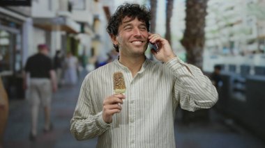 Güneşli bir günde, şehir dışında, açık havada telefonla konuşurken dondurmadan hoşlanan İspanyol bir adam çizgili gömlek giyiyor..