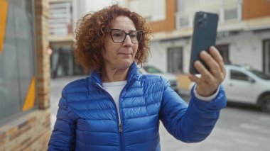 Şehir sokaklarında selfie çekerken gülümseyen, mavi ceketli ve gözlüklü bir kadın şehir ortamında akıllı telefonuyla bir anı yakalıyor..