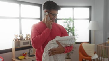 Yatak odasındaki genç adam telefonda konuşuyor. Elinde gözlük ve kırmızı kazak var. Etrafı oyuncaklarla ve mobilyalarla çevrili..