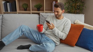 Akıllı telefonu ve kahvesi olan genç adam modern oturma odasındaki gri koltukta renkli yastıklar ve bitkilerle dinleniyor..
